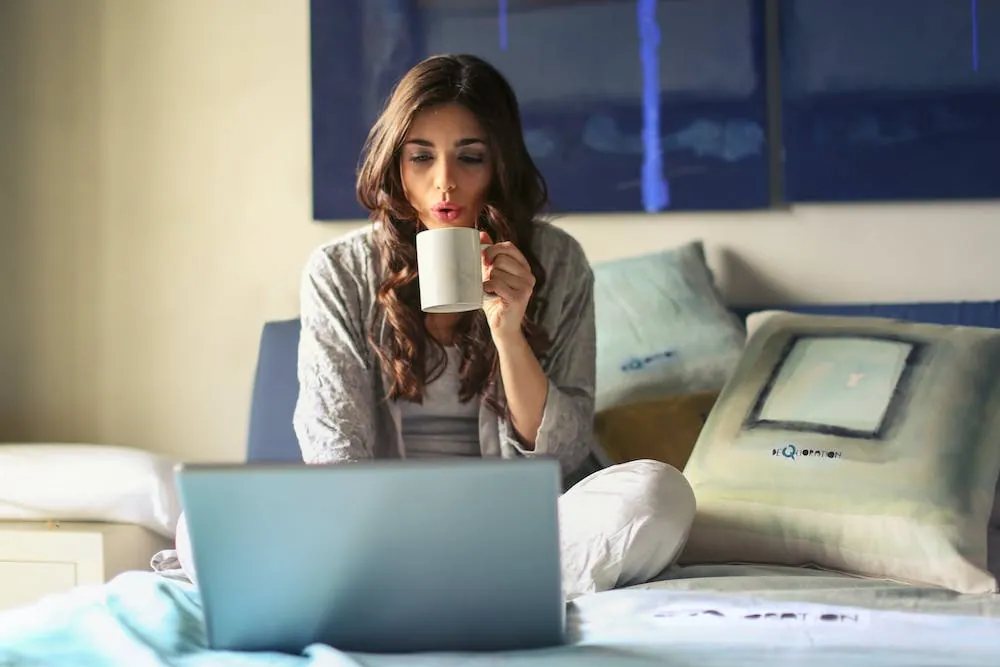 long brown hair woman drinking coffee on laptop sitting on bed